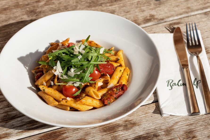 A plate of penne arrabbiata with arugula and parmesan on a wooden table, with a napkin showing the Balmalp logo.
