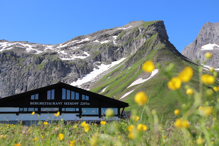 Das Seekopf Restaurant in 2207 m Seehöhe mit Berglandschaft im Hintergrund und gelben Blumen im Vordergrund.