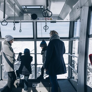 Four people in a cable car cabin with a view of snow-covered mountains.