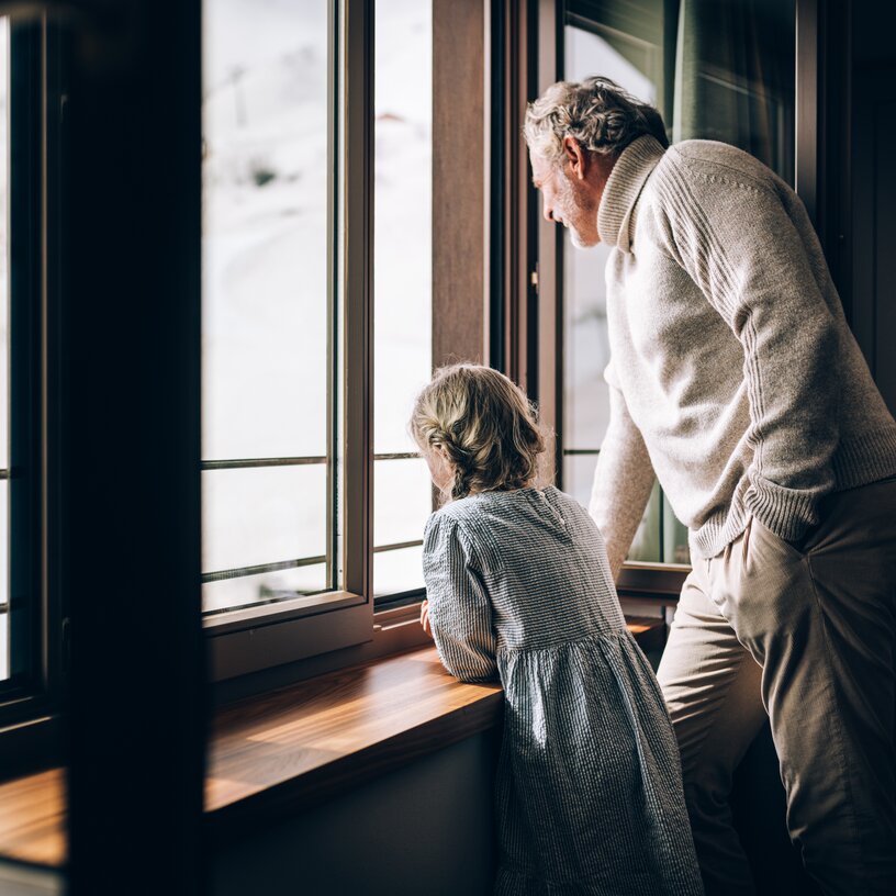 An older man and a young girl look out a window at snow-covered mountains.