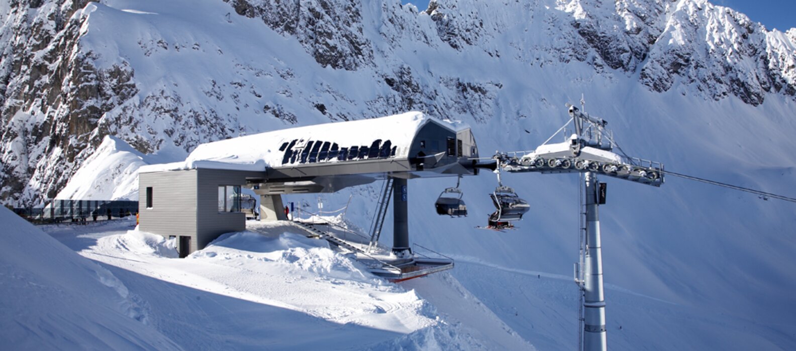 Schneebedeckte Bergstation mit Seilbahn und Bergen im Hintergrund.