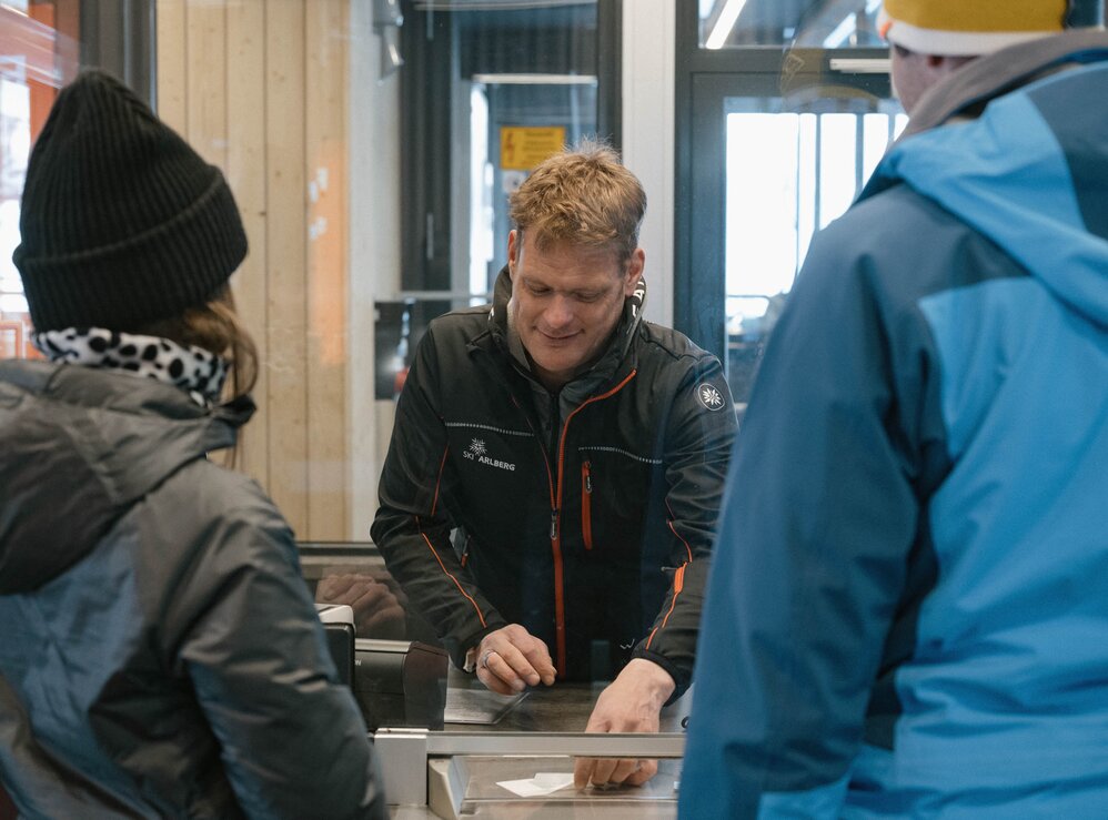 Menschen kaufen Tickets an einem Skischalter in einer Hütte.