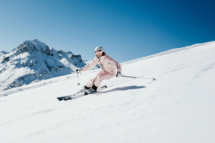 Person in rosafarbener Skiausrüstung fährt die Piste hinunter vor schneebedeckten Gipfeln.