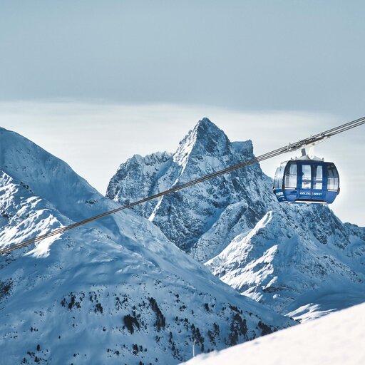 Blue cable car glides over a snowy mountain range with jagged peaks under a clear sky.