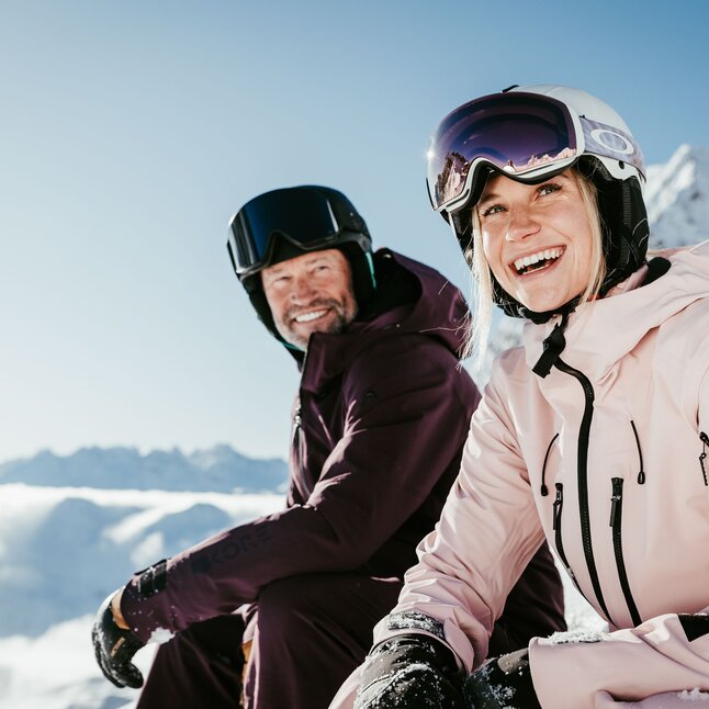 A couple in ski gear sitting and smiling in the snow.