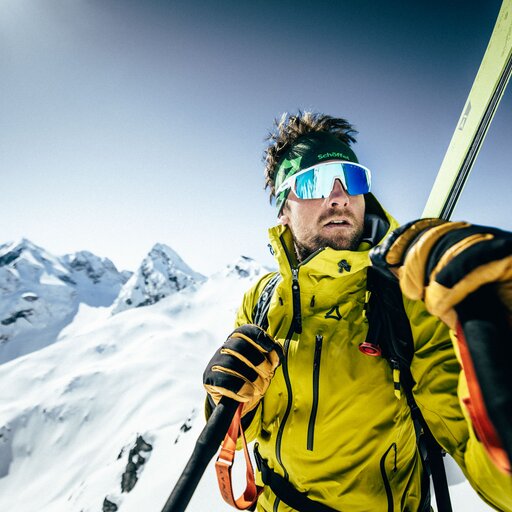 A skier wearing a yellow jacket and mirrored sunglasses holds ski poles on a snowy mountain range with sharp peaks.