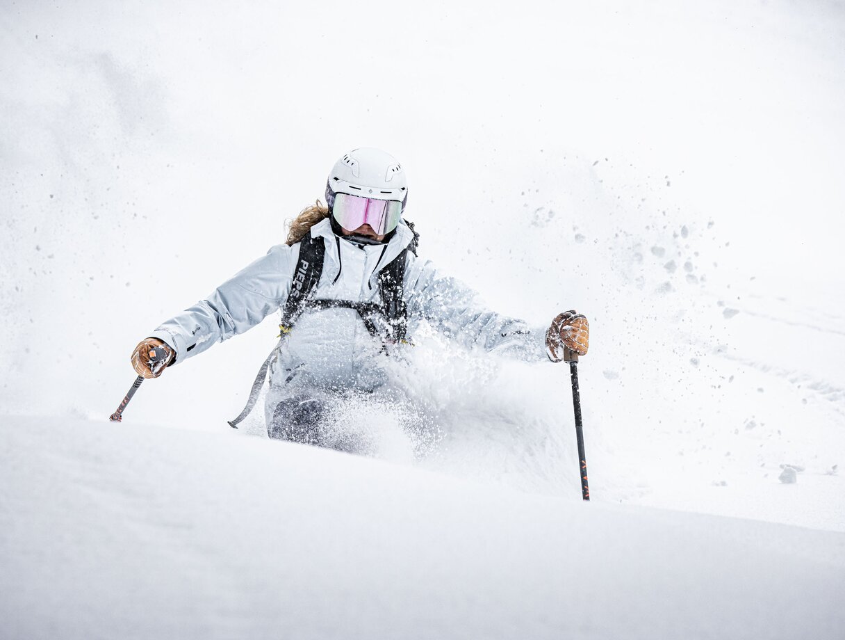 A skier in a white jacket and helmet carves through fresh powder, snow spray flying around them.