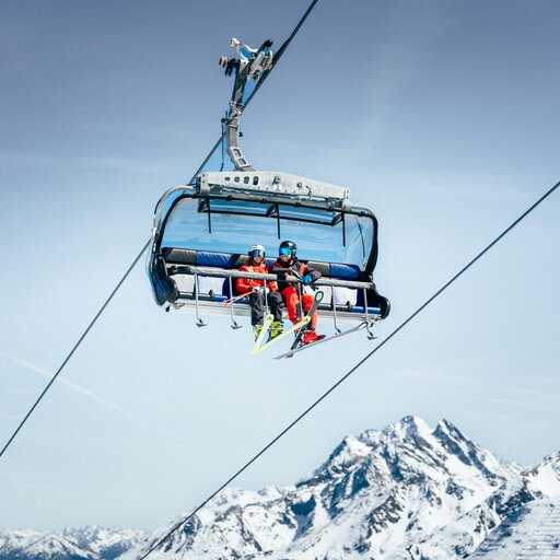 Two skiers sit on a chairlift, wearing helmets and colorful gear, with snow-covered mountains in the background.