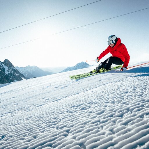 A skier in a red jacket carving down a snowy slope, with mountain peaks in the background.