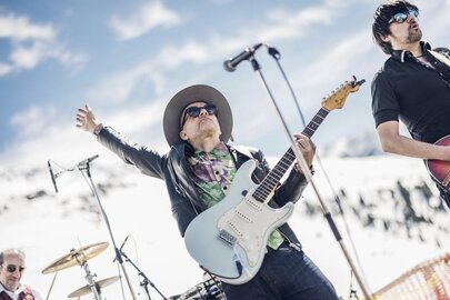 Band spielt mit Gitarren und Schlagzeug vor schneebedeckten Bergen.