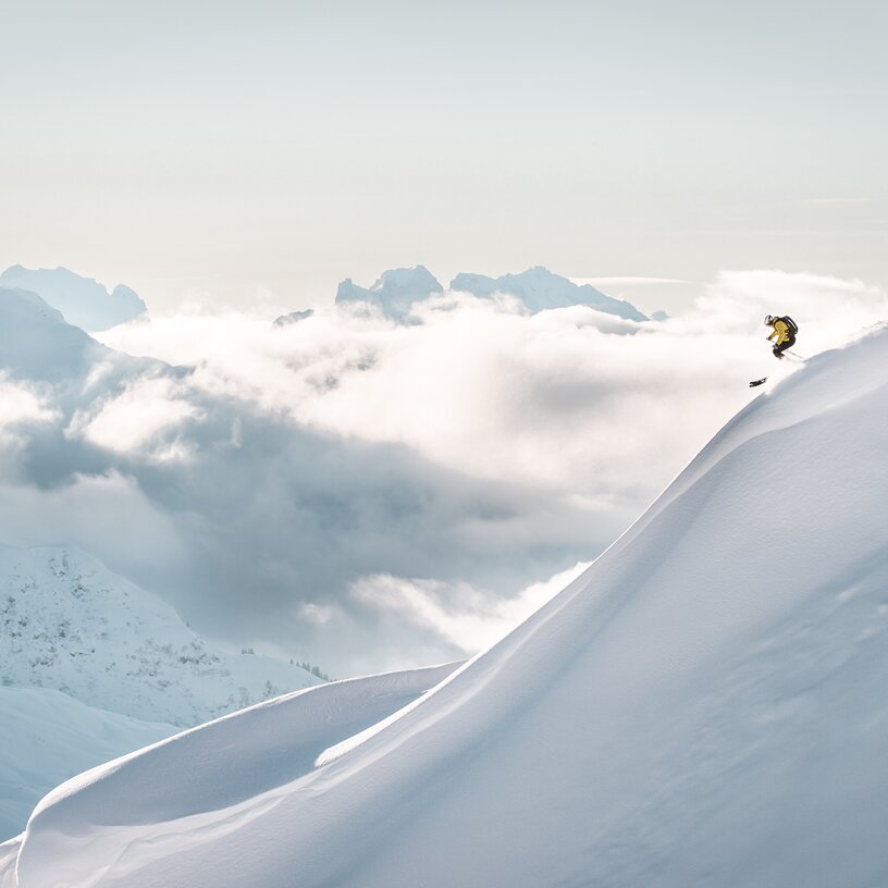 A skier in a yellow jacket descends a snowy slope with clouds and distant mountain peaks.