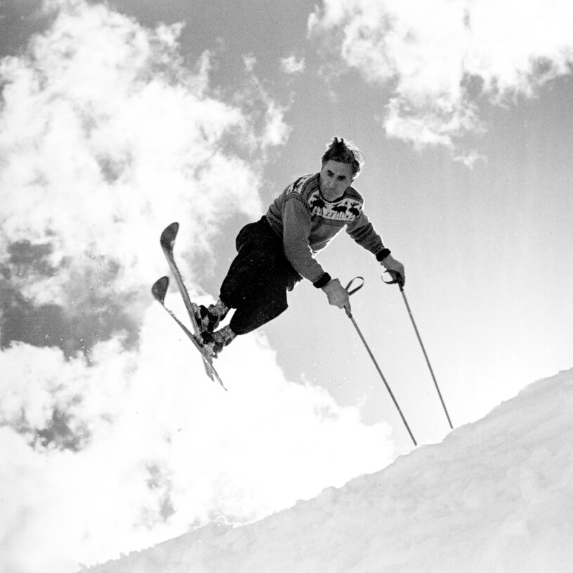 Black-and-white photo of a skier mid-air jumping off a snowy slope, with skis in the air against a cloudy sky.