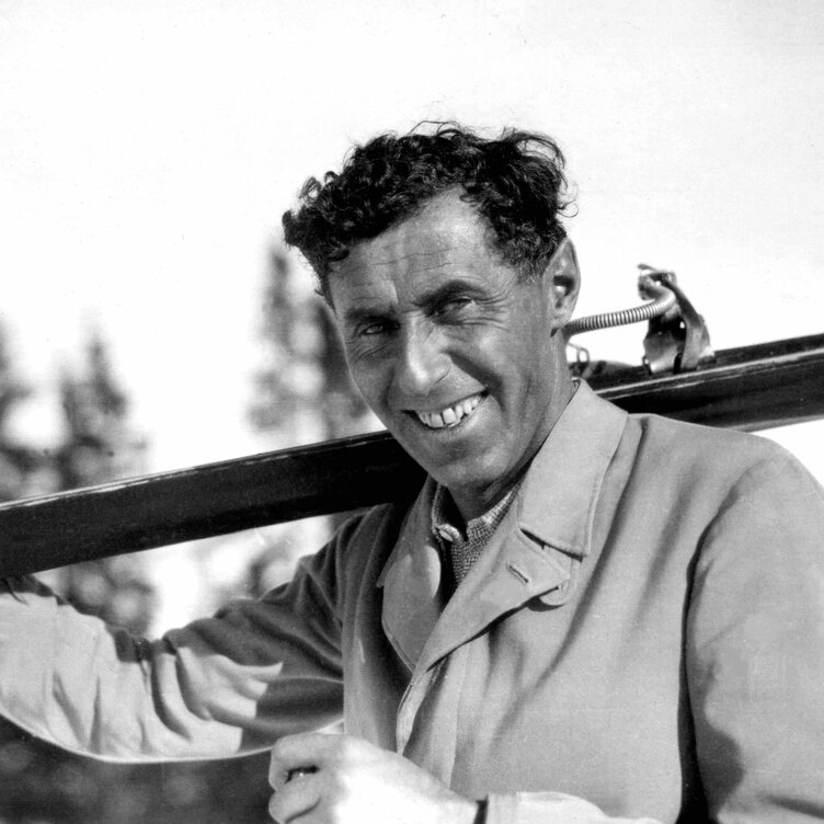 A man smiling while carrying skis on his shoulder outdoors.
