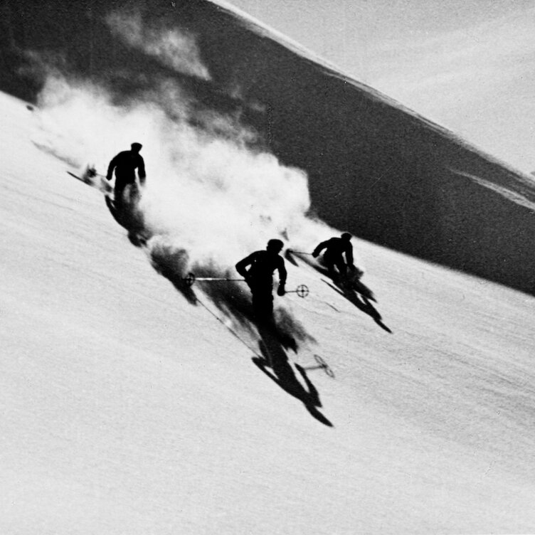 Three skiers race down a snowy slope, creating clouds of snow and dramatic shadows in the afternoon sun.