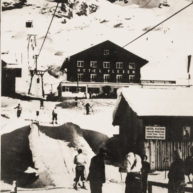 Historic image of skiers in the snow in front of Hotel Flexen, with a cable car in the background.