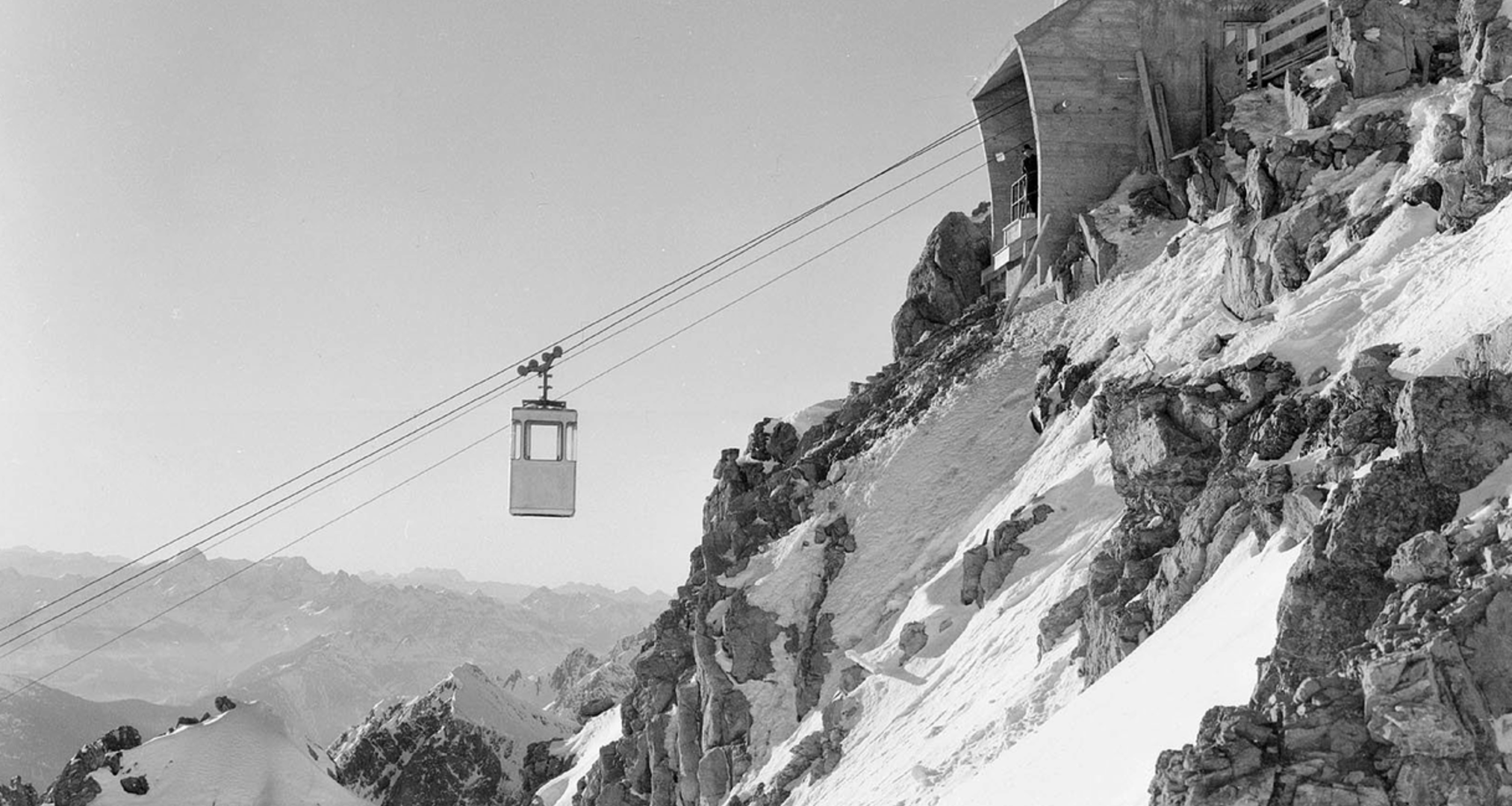 Black-and-white image of a cable car on a snow-covered mountain peak.