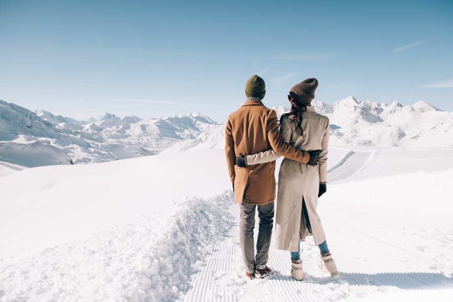 Ein Paar in Winterkleidung steht umarmt vor einer Berglandschaft im Schnee.