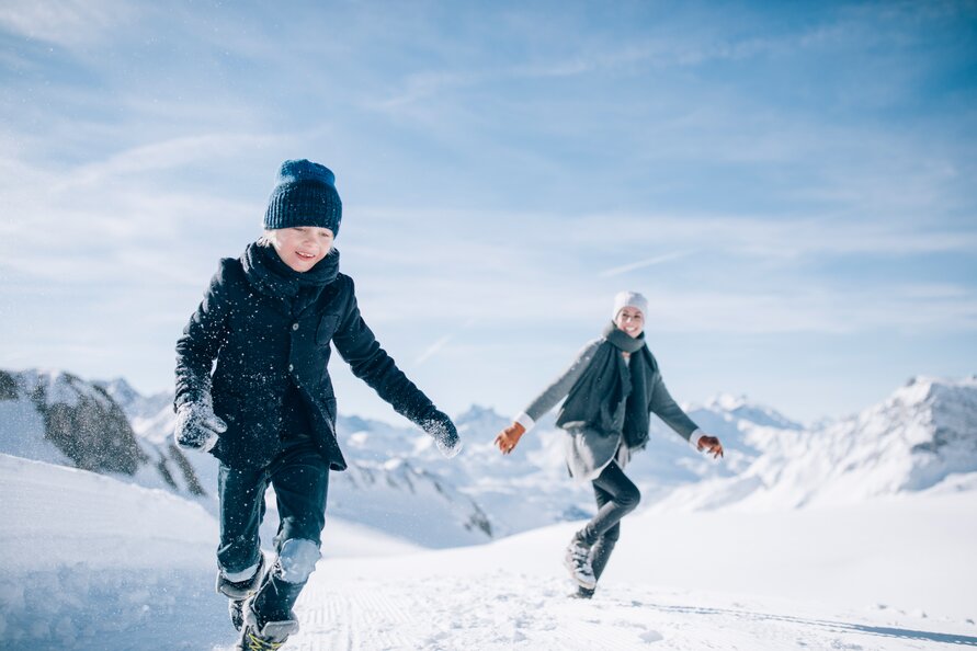 Mutter spielt mit ihrem Sohn im Schnee bei sonnigem Wetter und Bergen im Hintergrund.