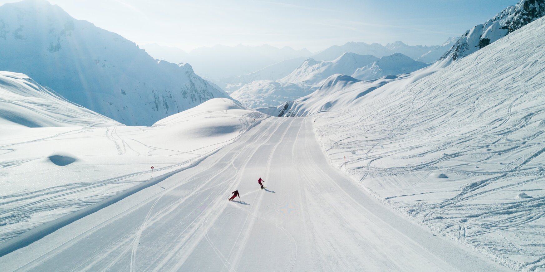 Zwei Skifahrer fahren auf breiter, schneebedeckter Piste.