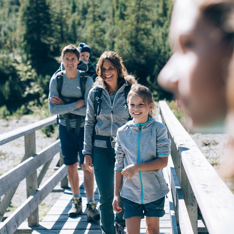 Familie wandert auf Holzbrücke über Fluss mit Wald im Hintergrund.