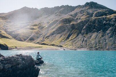 Person sitting on a rock by a clear mountain lake with green hills and mountains in the background.