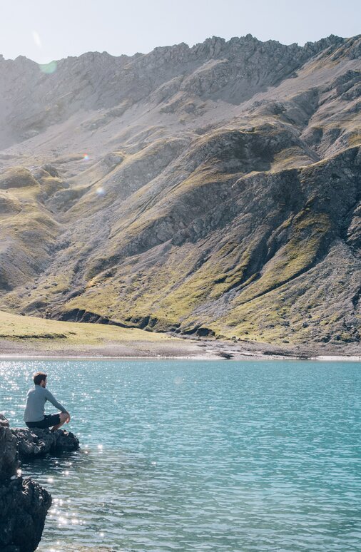 Person sitting on a rock by a clear mountain lake with green hills and mountains in the background.