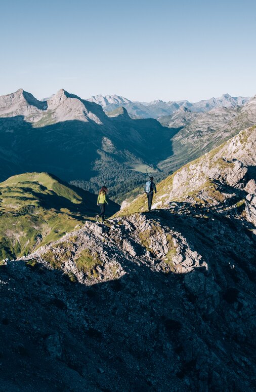 Two people hiking on a mountain ridge with panoramic views of the Alps.