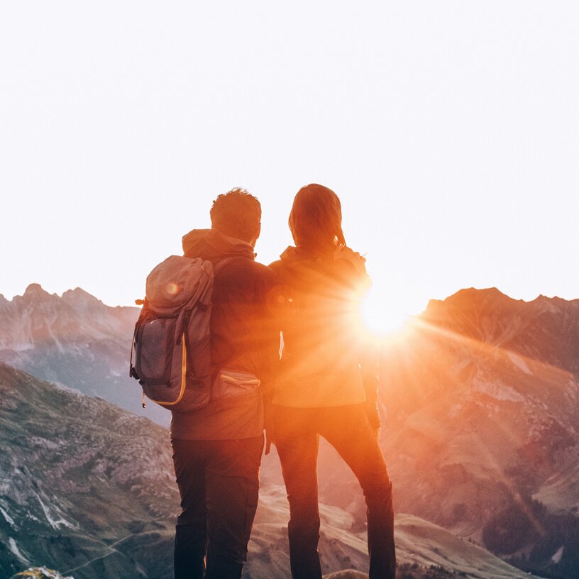 Zwei Personen stehen auf einem Berg und schauen in den Sonnenaufgang.