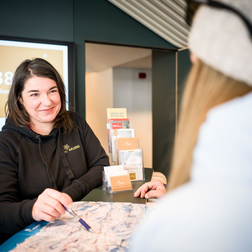 Eine Frau im Infopoint erklärt einer Skifahrerin mit einer Karte das Skigebiet Ski Arlberg.