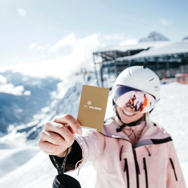 Lächelnde Skifahrerin hält goldenen Skipass von Ski Arlberg ausgestreckt vor sich.