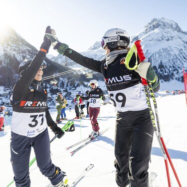 Zwei Skifahrer geben sich nach einem Skirennen ein High-Five. Mehrere Teilnehmer und schneebedeckte Berge sind im Hintergrund zu sehen.
