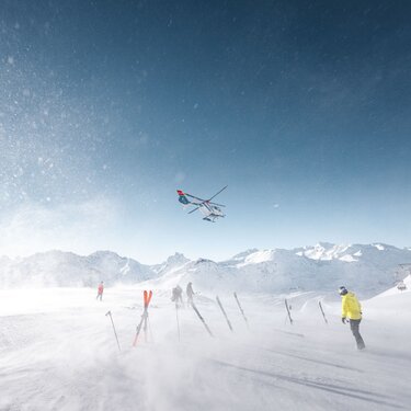 Ein Helikopter fliegt über Skifahrer in einer verschneiten Bergregion.