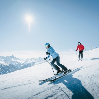 Two people skiing on a sunny slope with mountains in the background.