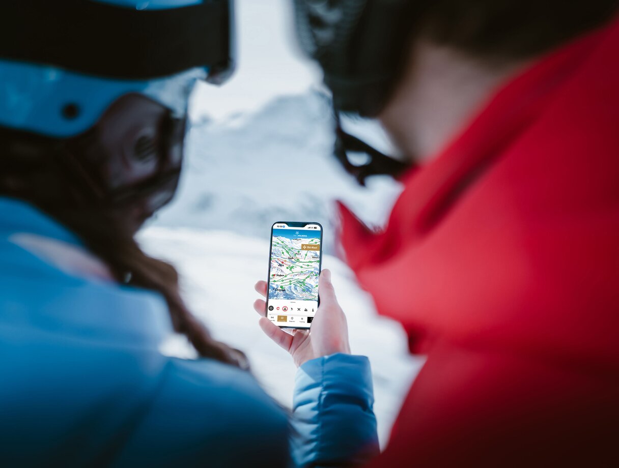 Zwei Personen überprüfen Skiroute auf einem Smartphone mit schneebedeckten Bergen im Hintergrund.