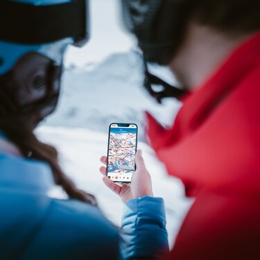 Two people check ski route on a smartphone with snowy mountains in the background.