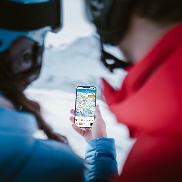 Zwei Personen überprüfen Skiroute auf einem Smartphone mit schneebedeckten Bergen im Hintergrund.