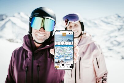 Skipärchen zeigt eine Mobil-App bei sonniger Berglandschaft.
