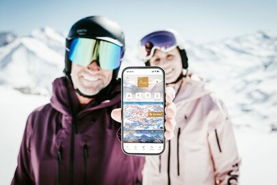 Skipärchen zeigt eine Mobil-App bei sonniger Berglandschaft.
