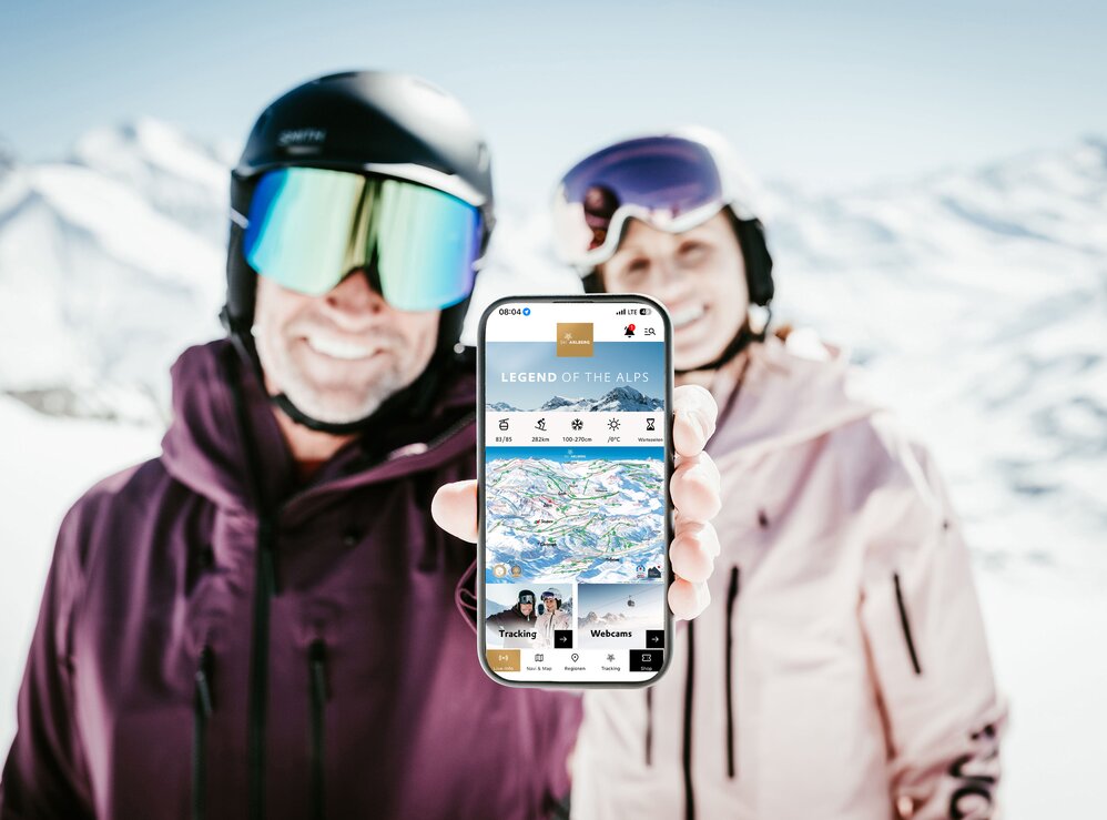 Ski couple showing mobile app in sunny mountain landscape.
