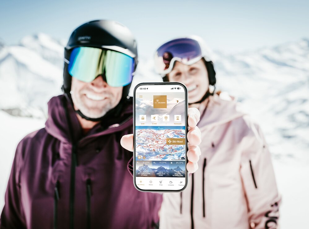 Ski couple showing mobile app in sunny mountain landscape.