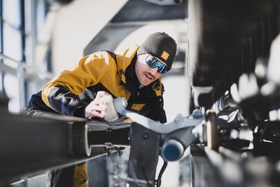 Techniker in einer gelb-schwarzen Jacke, einer schwarzen Mütze und Sonnenbrille inspiziert eine Maschine in der Talstation.