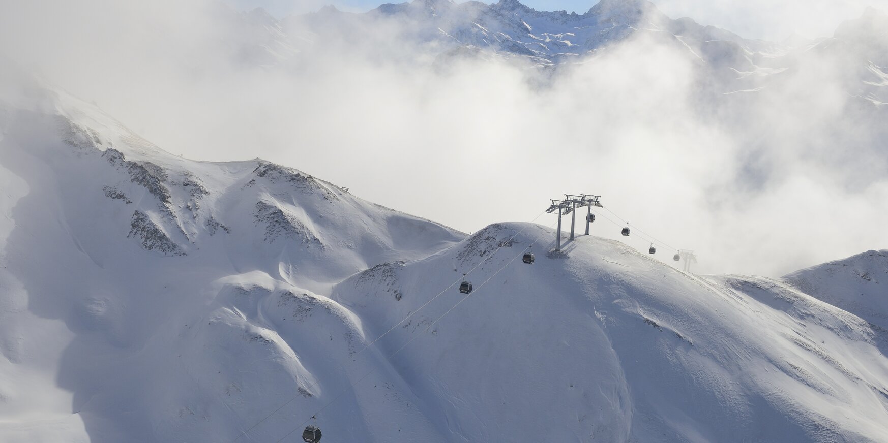 Snow-covered mountain landscape with a ski lift and chairlifts, clouds rolling over the peaks.
