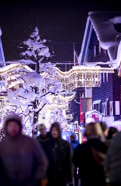 Menschen spazieren durch eine verschneite, beleuchtete Straße.