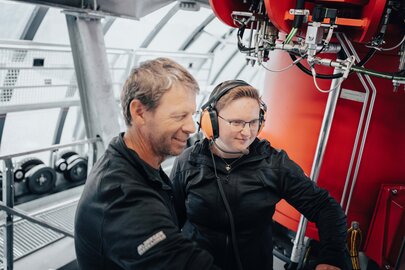 Zwei Personen arbeiten an einer großen roten Maschine mit Kopfhörern.