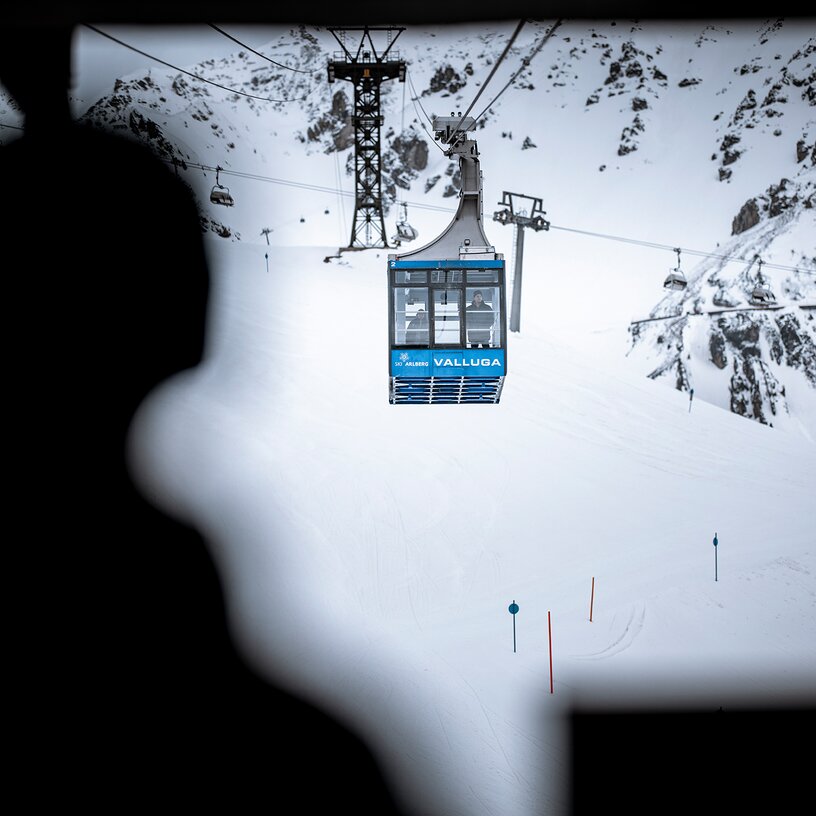 Eine Personensilhouette steht am Eingang zur Einfahrt der Vallugabahn und schaut hinaus zu einer Seilbahn. Rundherum sind verschneite Berge.