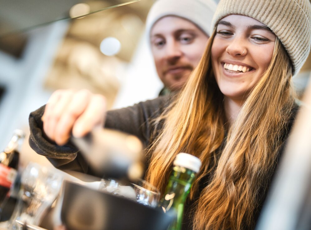 A smiling couple gets something to eat and drink at the buffet in a ski lodge. They are wearing warm clothes and hats.    