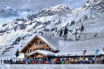 Verschneite Almhütte mit Menschen auf der Terrasse und Bergkulisse im Hintergrund.