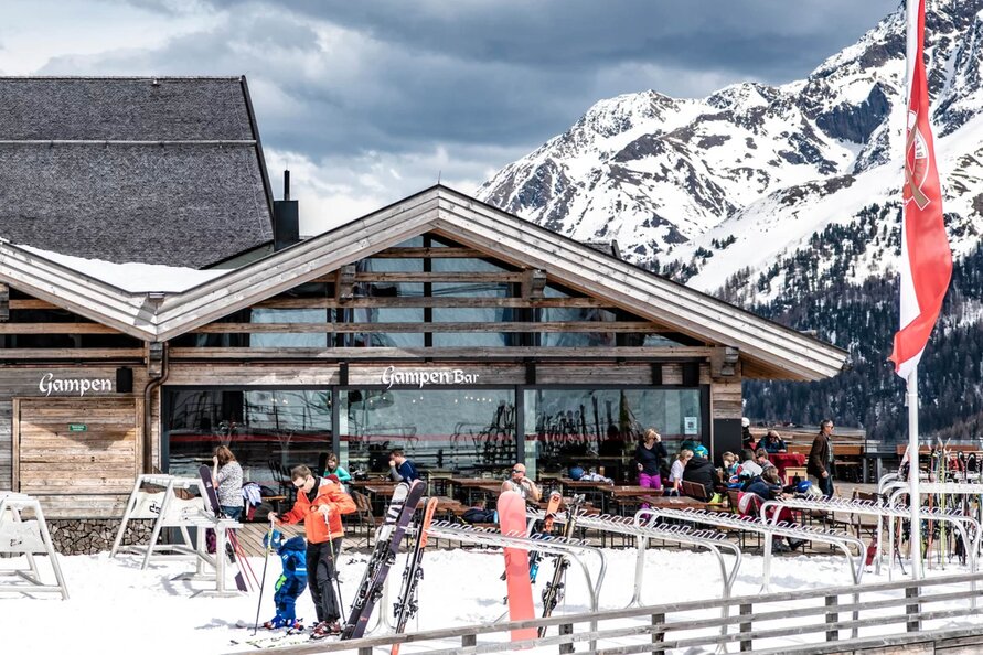 Skiers outside Gampen Restaurant with snowy mountains in the background.