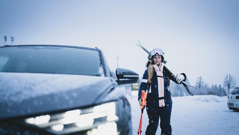 Woman in ski gear walking with skis and poles next to a car in the snow.