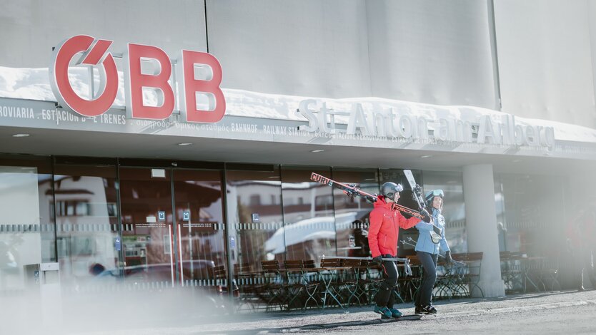 Zwei Skifahrer in Skikleidung verlassen den Bahnhof St. Anton am Arlberg mit Skiern auf den Schultern.